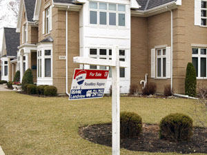 A "For Sale" sign swings in a breeze in front of a 