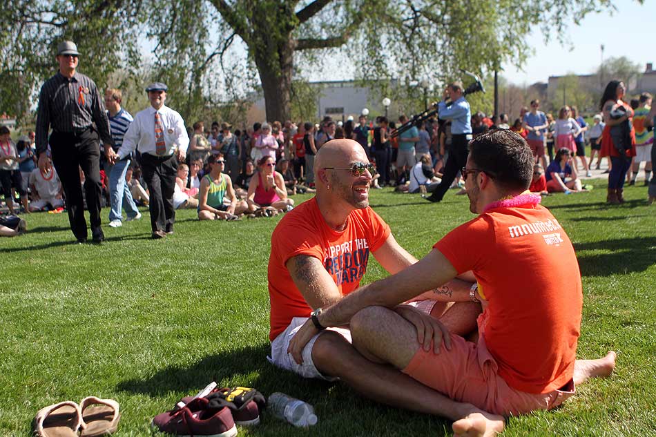 Two men sit close together on the grass, facing each other, hands on each other's thighs, with a little bit of space between them and the standing crowds around them, a lovely intimate moment in such a public place.